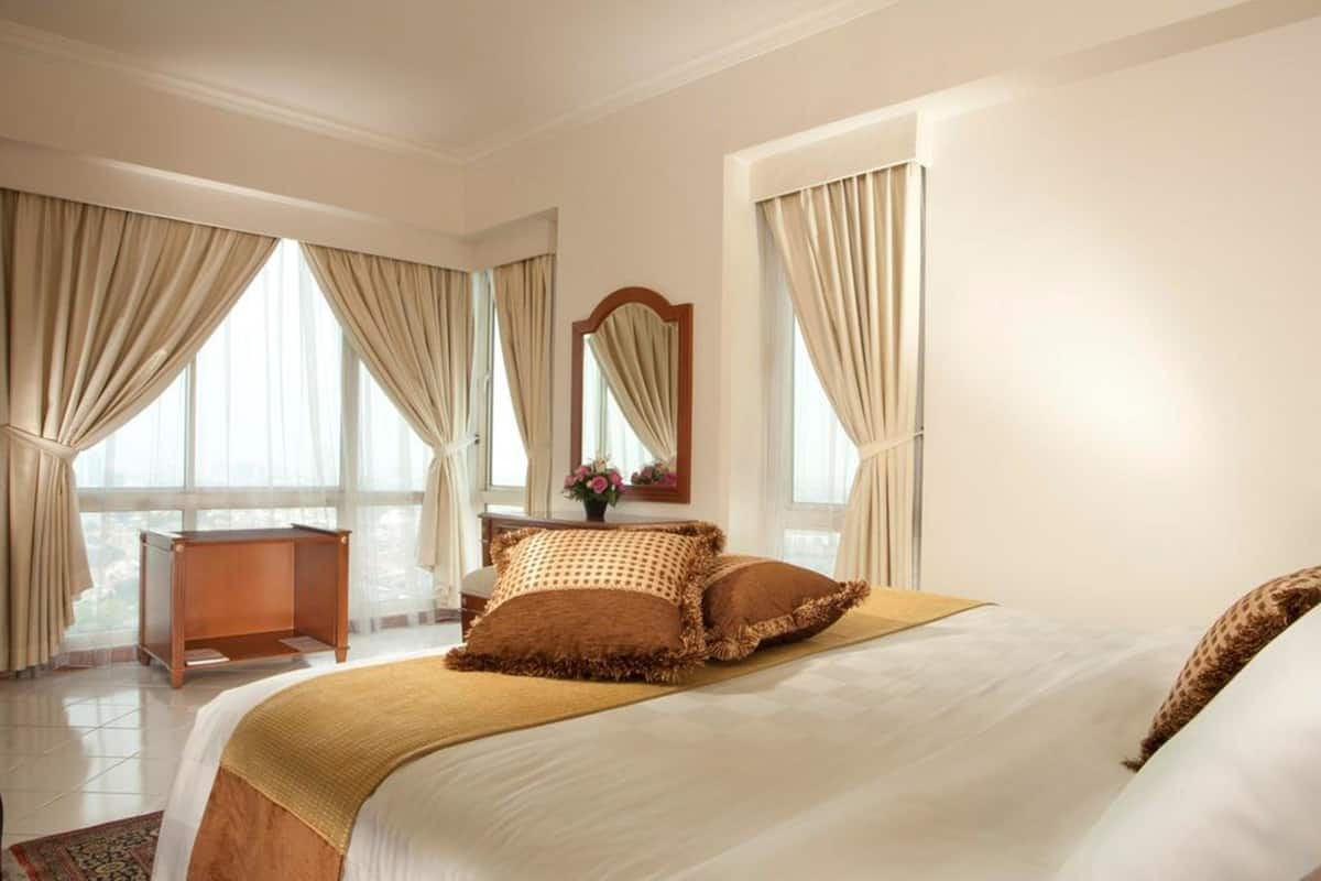 Ein Hotelzimmer mit einem großen Bett, einem Nachttisch aus Holz, einem Spiegel und Blick auf die Stadt durch große Fenster mit beigen Vorhängen.