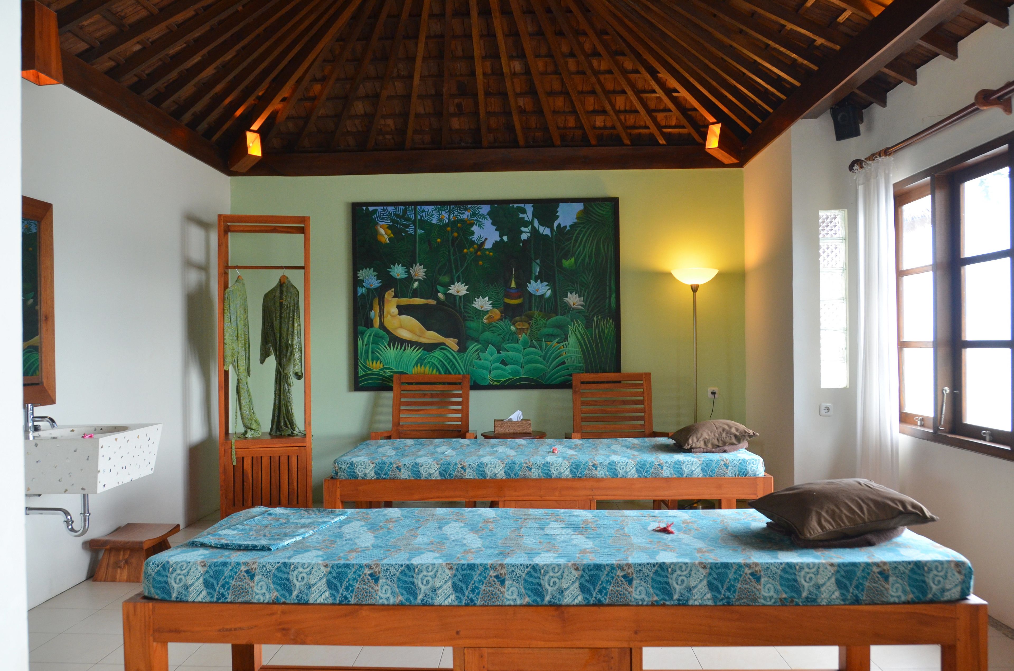 Une pièce avec deux lits en bois, un tableau représentant un paysage tropical, un lavabo et une fenêtre avec des rideaux.