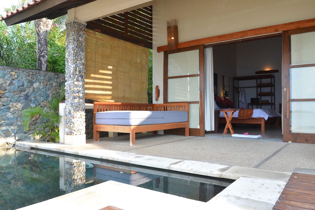 A poolside area with a wooden bench, a stone wall, and a view into a room with a bed and a table.