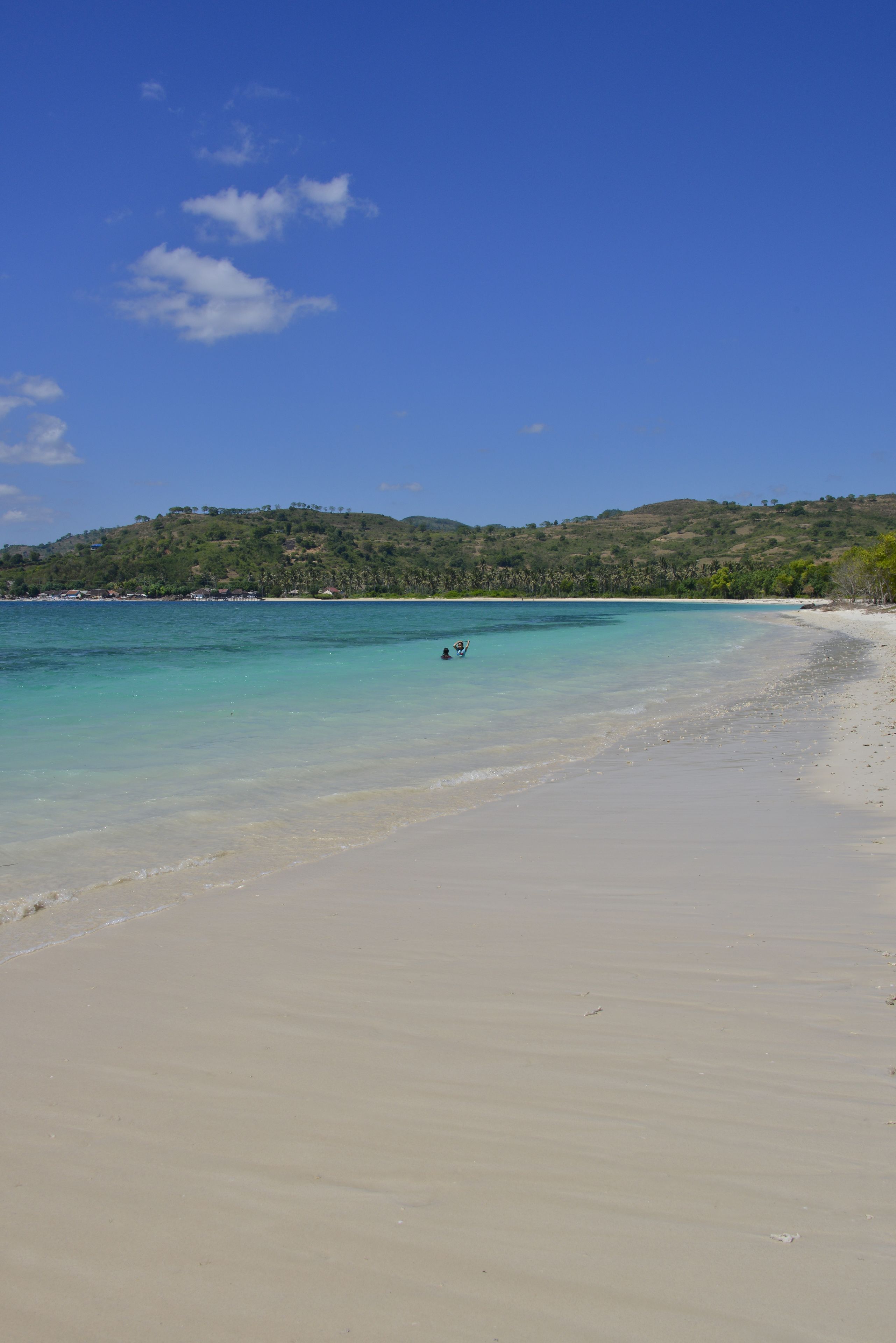 Une plage à l’eau turquoise et limpide, un rivage sablonneux et un paysage vallonné en arrière-plan.