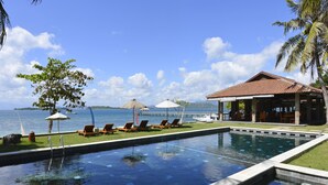 A pool with lounge chairs and umbrellas, a building with a red roof, and a view of the sea. - Cocotinos Sekotong, a Boutique Beach Resort & Spa (Sekotong Barat)