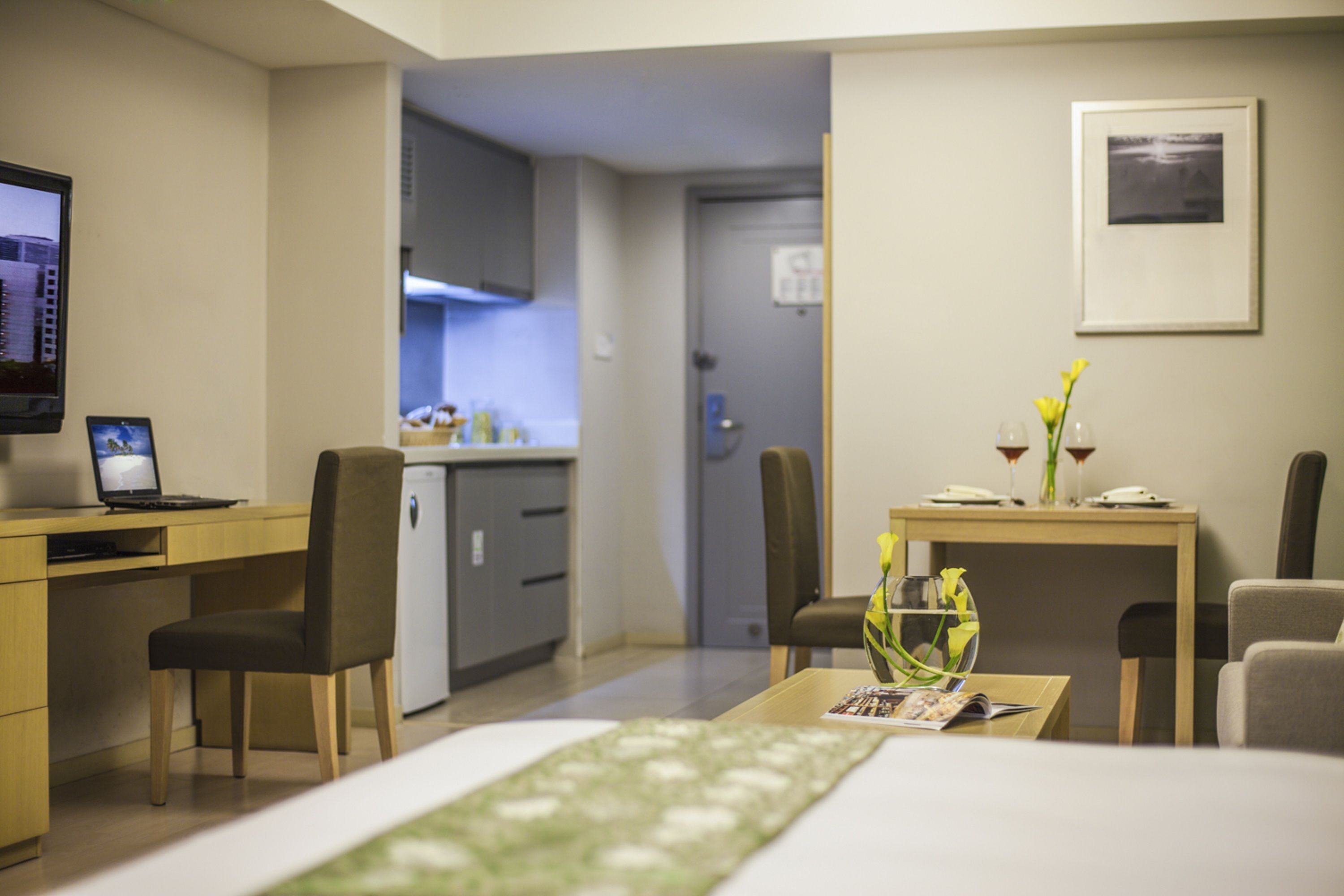 Habitación de hotel con cama, escritorio con computadora portátil, silla, pequeña cocina y mesa de comedor con un florero.