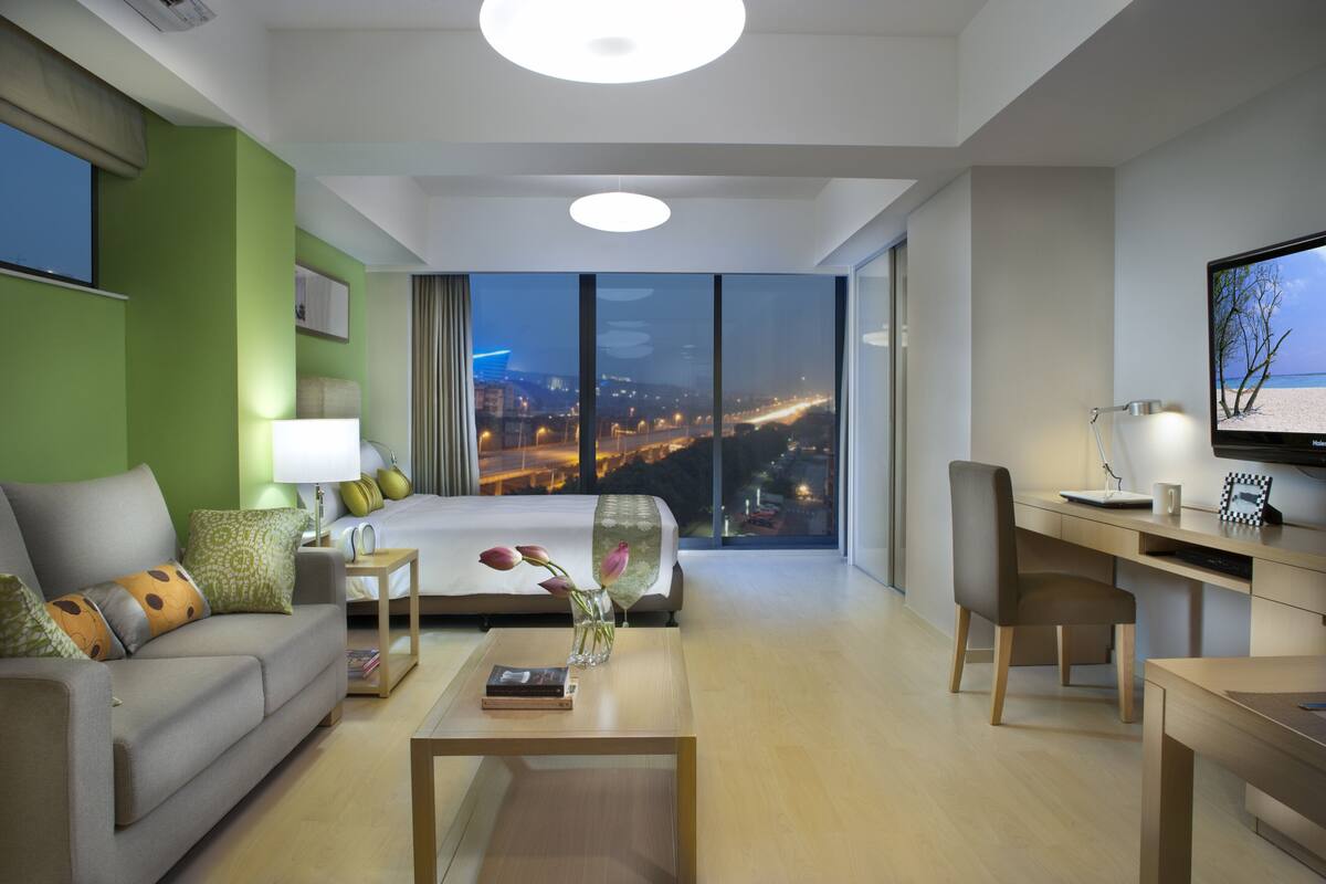 A modern hotel room with a large bed, a desk, a sofa, and a view of the city at night.