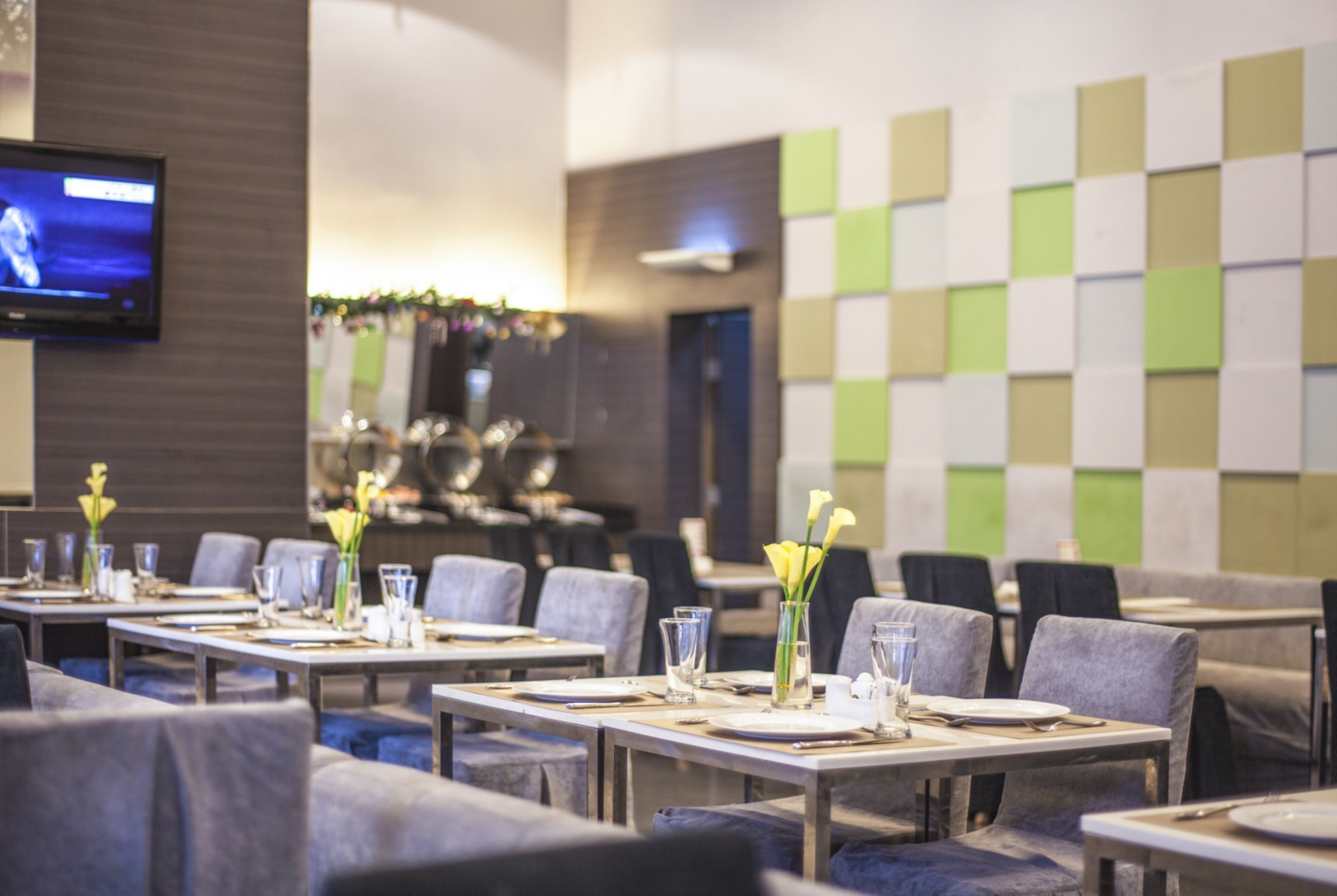 A restaurant with tables set for dining, a television screen, and a wall with a checkered pattern.