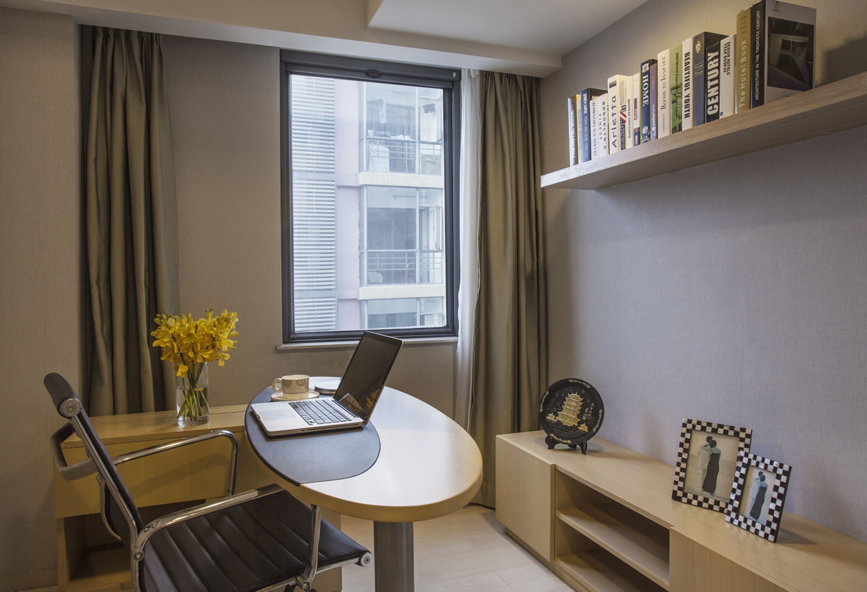 A modern room with a laptop on a round table, a chair, a vase with yellow flowers, a window with blinds, and a bookshelf with books.