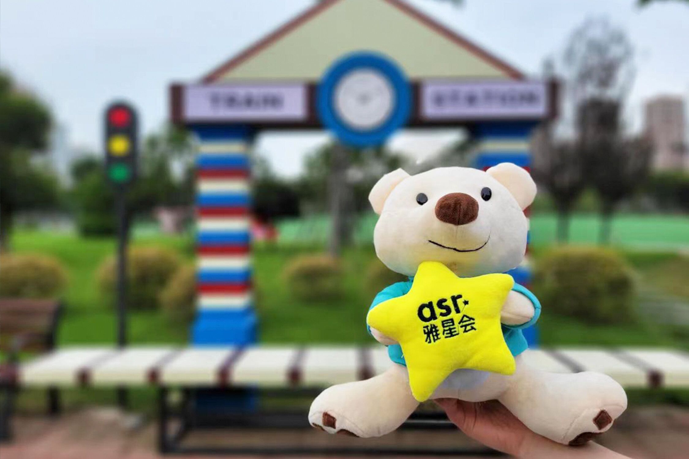 A plush toy bear holding a yellow star-shaped object with text, in front of a building with a clock and signage.