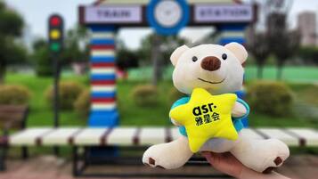 A plush toy bear holding a yellow star-shaped object with text, in front of a building with a clock and signage.