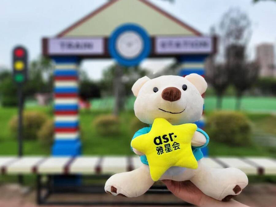 A plush toy bear holding a yellow star-shaped object with text, in front of a building with a clock and signage.