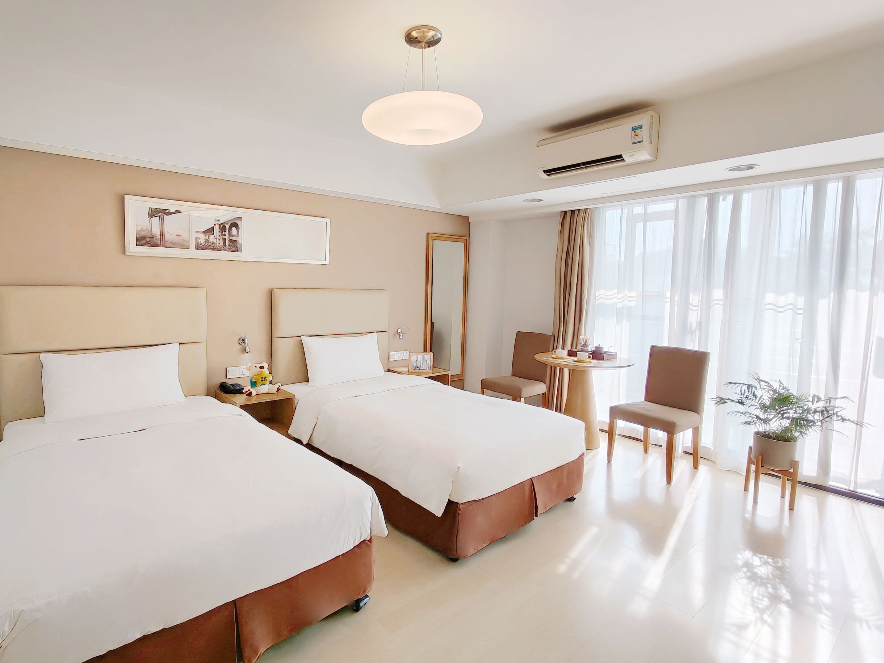 A hotel room with two beds, a small table with a chair, a potted plant, and a large window with curtains.
