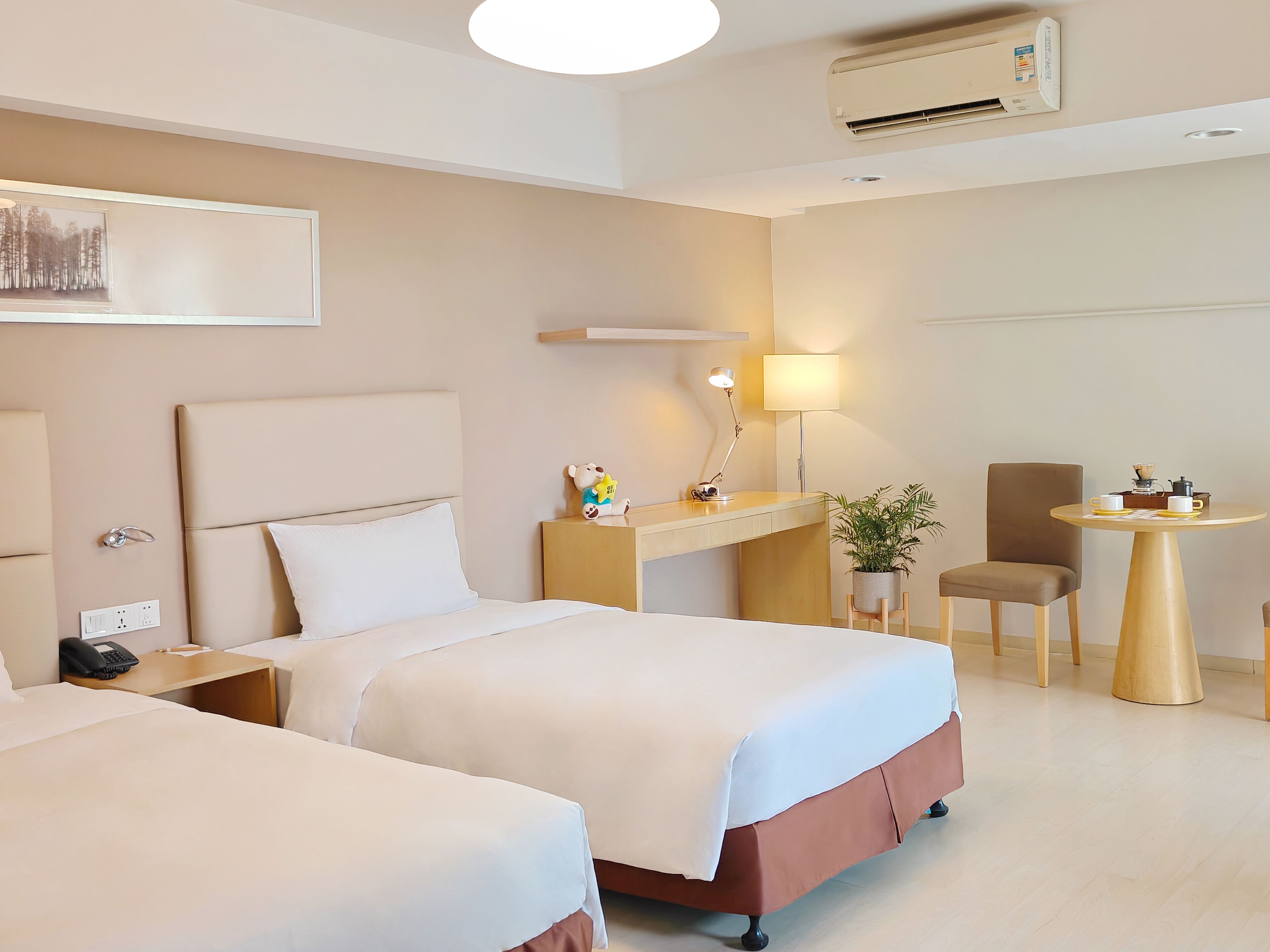 A hotel room with two beds, a desk, a chair, a lamp, a small table with a plant, and an air conditioning unit.