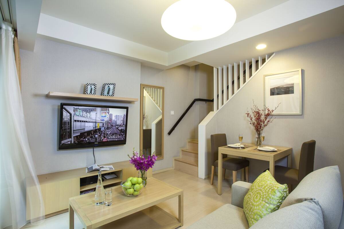 A modern living room with a flat-screen TV, a wooden coffee table with decorative items, a dining area with a table and chairs, and a staircase with a railing.