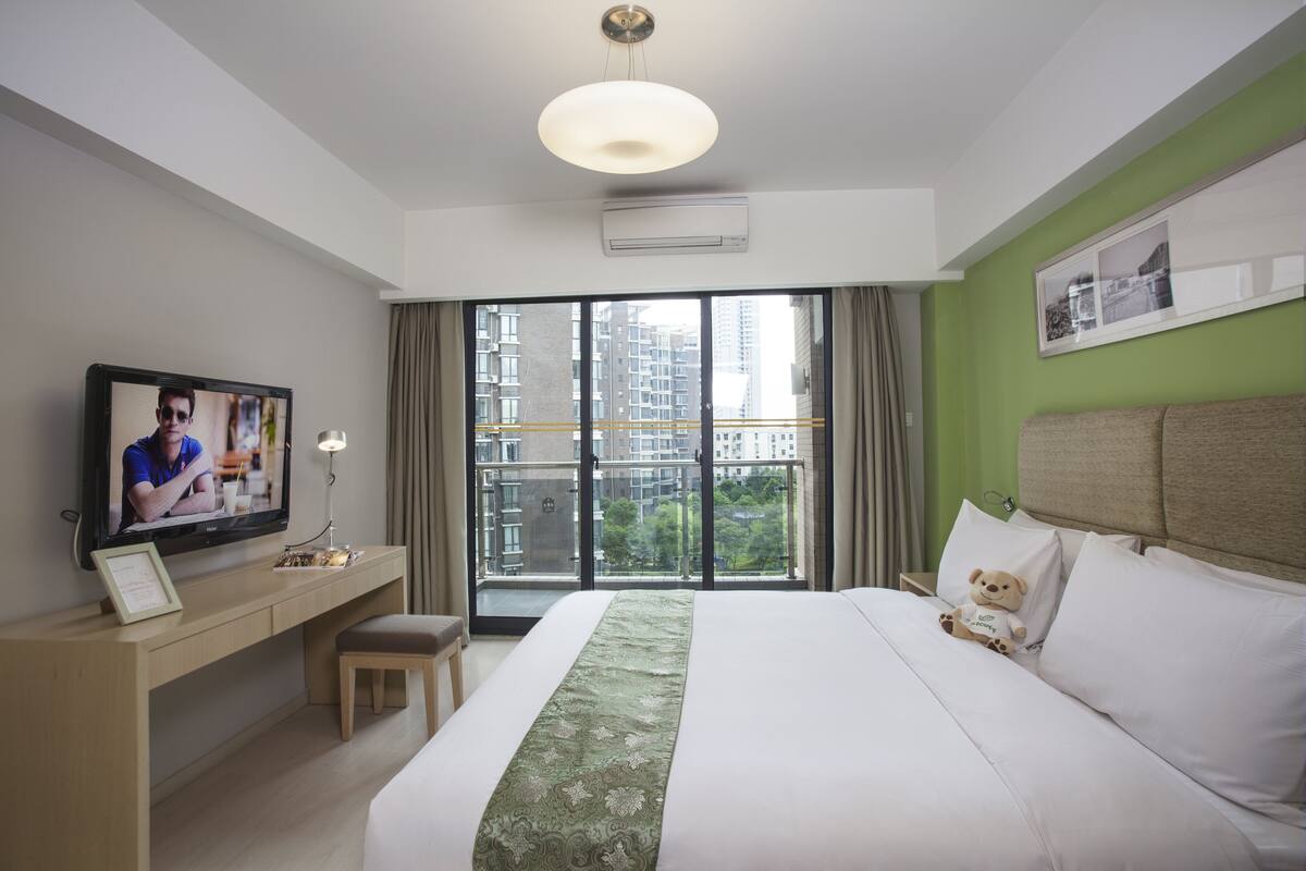 A hotel room with a large bed, a desk with a television, a teddy bear on the bed, and a balcony with a view of buildings.