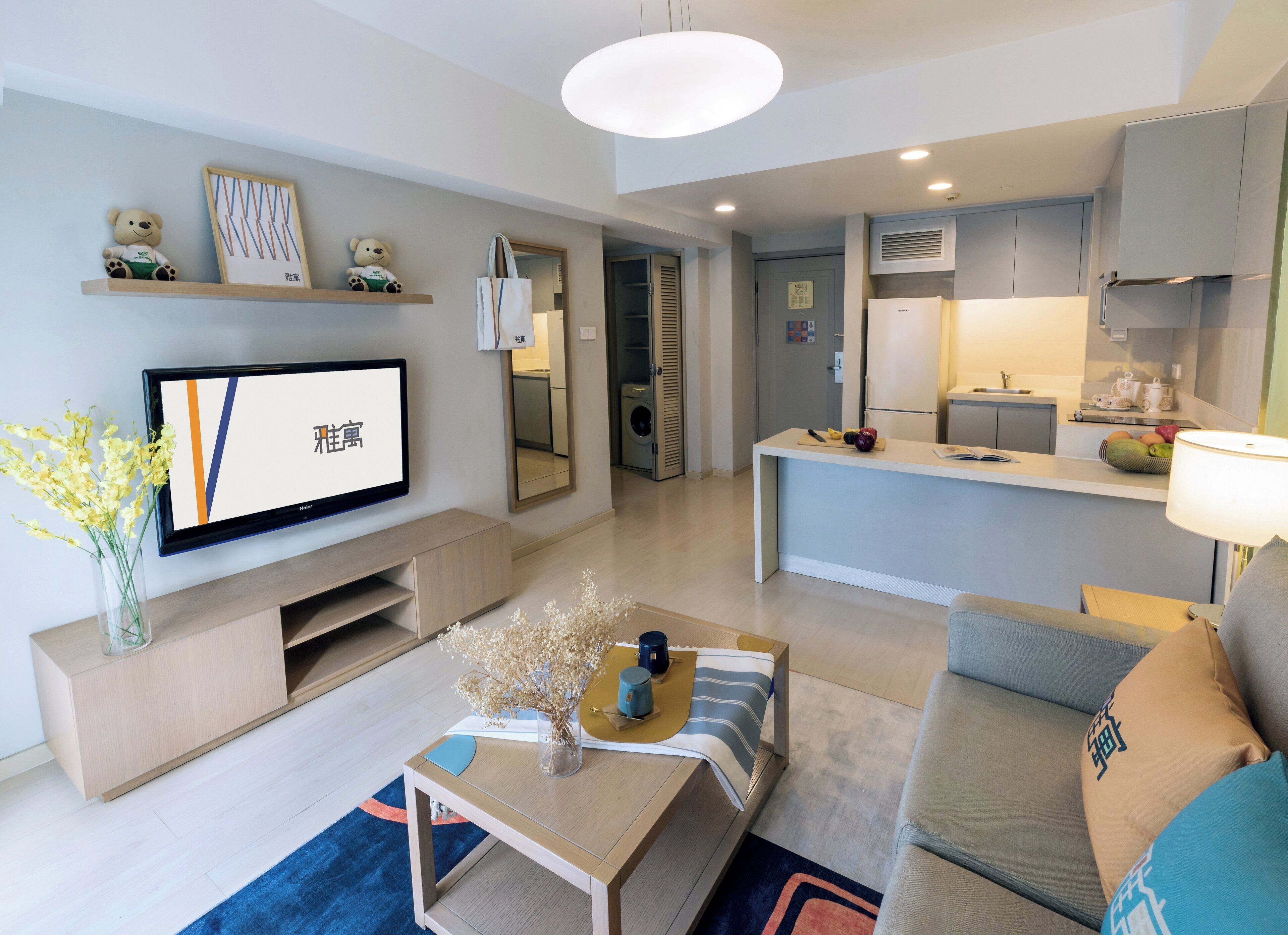 A modern living room with a flat-screen TV, a coffee table with decorative items, a sofa, and a kitchen area with a refrigerator and microwave.
