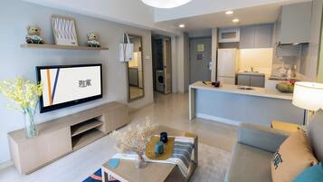 A modern living room with a flat-screen TV, a coffee table with decorative items, a sofa, and a kitchen area with a refrigerator and microwave.
