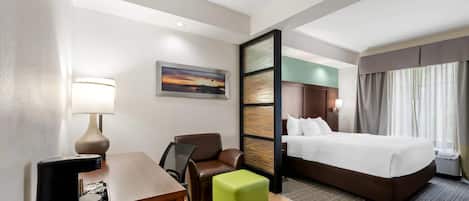 Una habitación de hotel con una cama, un escritorio con lámpara, una silla y un pequeño reposapiés.