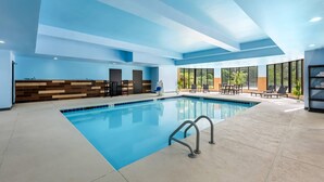 An indoor swimming pool with a clear blue pool, a poolside area with chairs and tables, and large windows offering a view of greenery outside. - Comfort Suites New Bern near Cherry Point (New Bern)