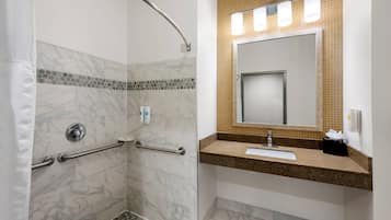 A bathroom with a shower, grab bars, a sink with a mirror, and a towel dispenser.