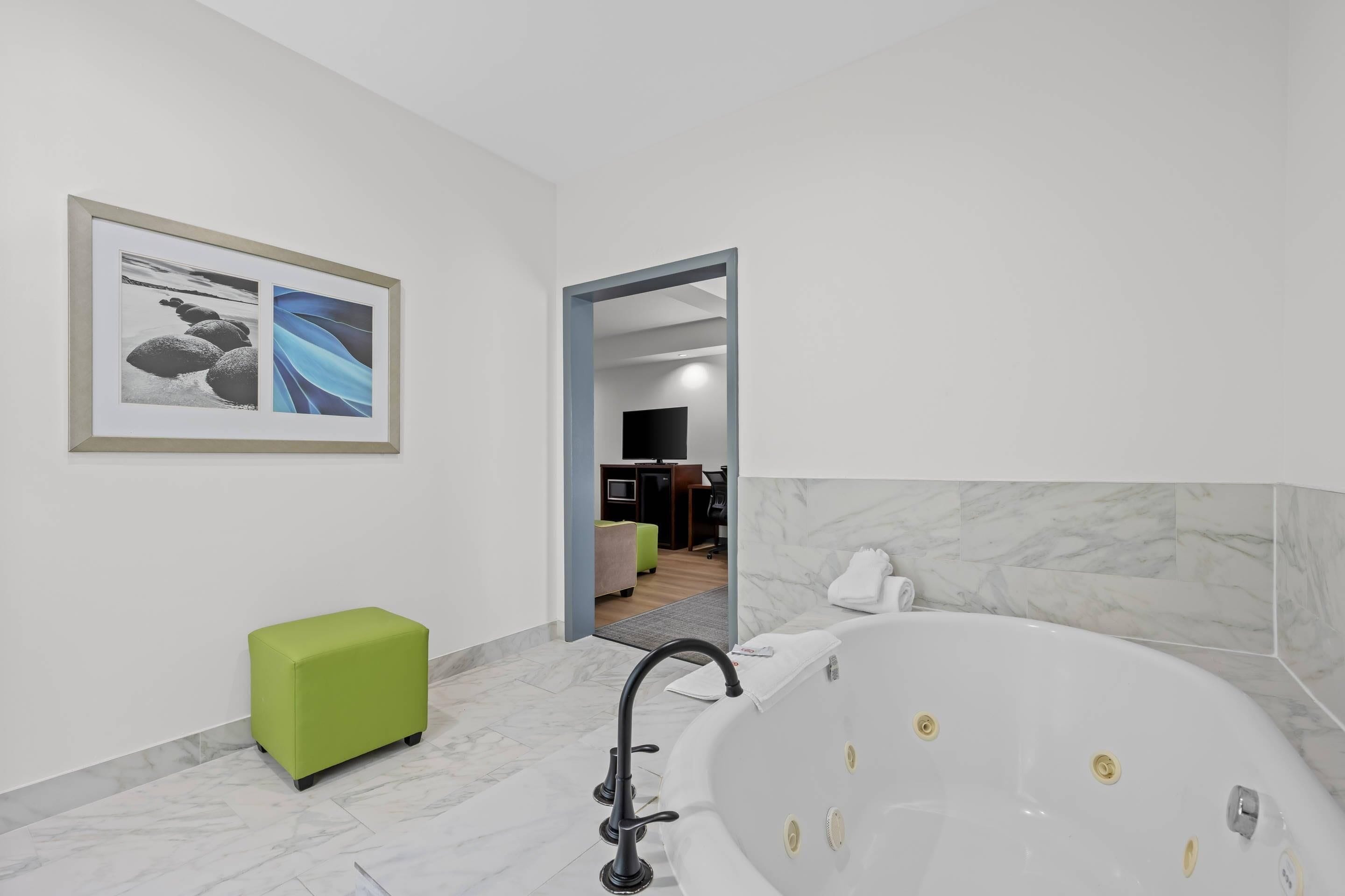 Een moderne badkamer met een marmeren vloer, een groene poef, een groot bad en een ingelijst kunstwerk aan de muur.