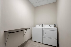 Une buanderie équipée de deux machines à laver et deux sèche-linge superposés, d’une étagère en métal noir et d’un sol carrelé.