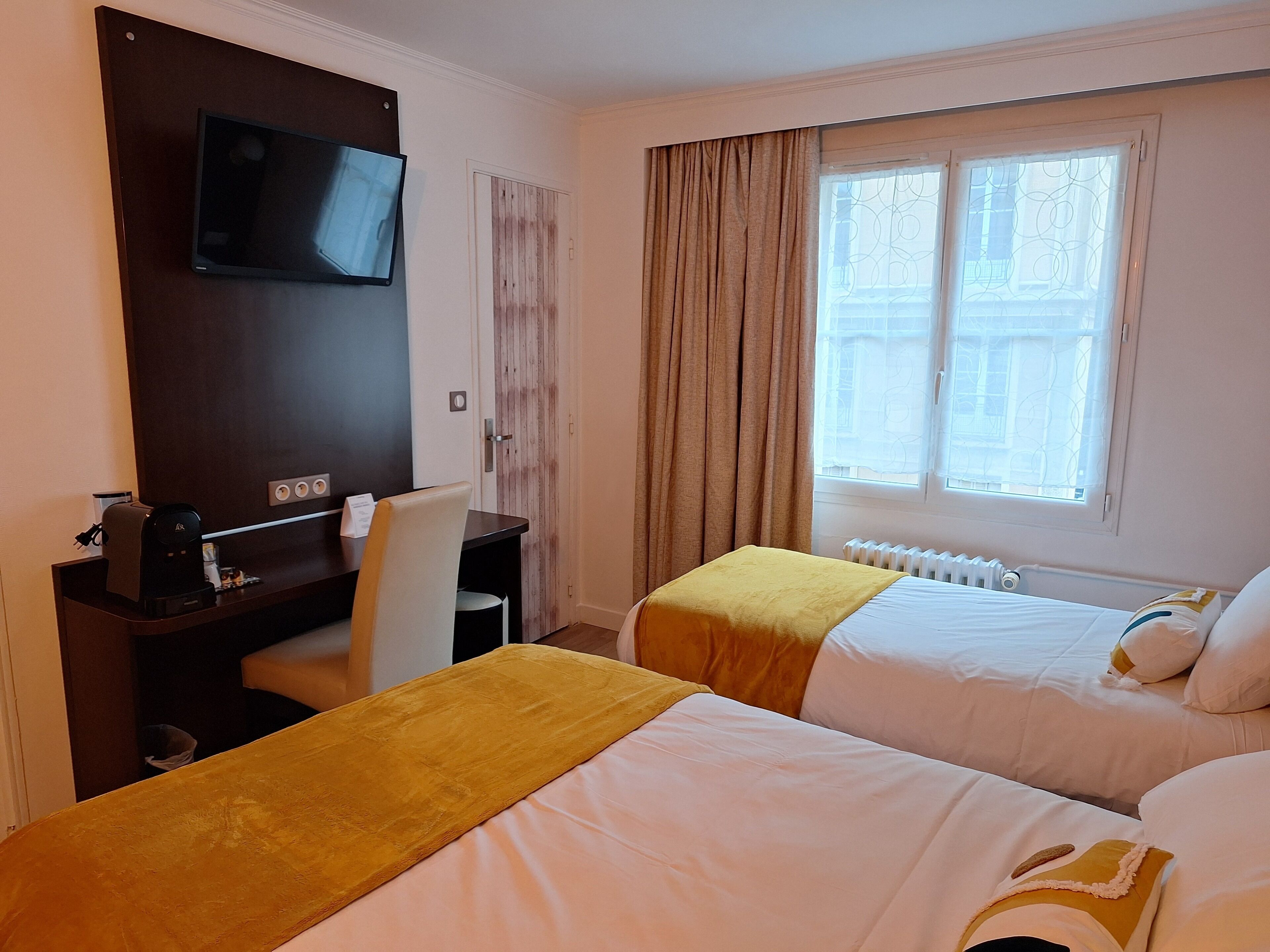 A hotel room with two beds, a desk with a coffee maker, a TV mounted on the wall, and a window with curtains.