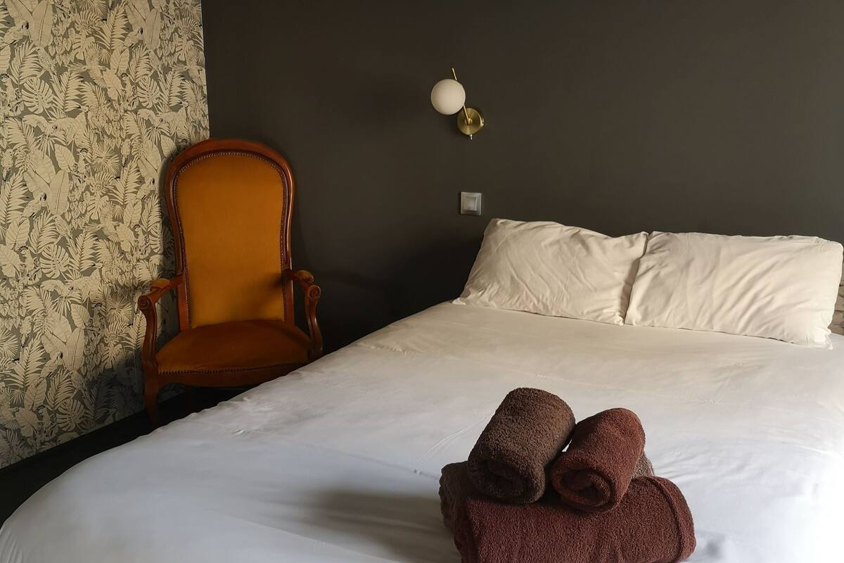 Una habitación de hotel con una cama grande, una silla de madera, una lámpara de pared y un papel tapiz estampado con motivos florales.