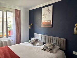 A bedroom with a bed, a window with curtains, a wall-mounted light, and a painting of palm trees and a van.