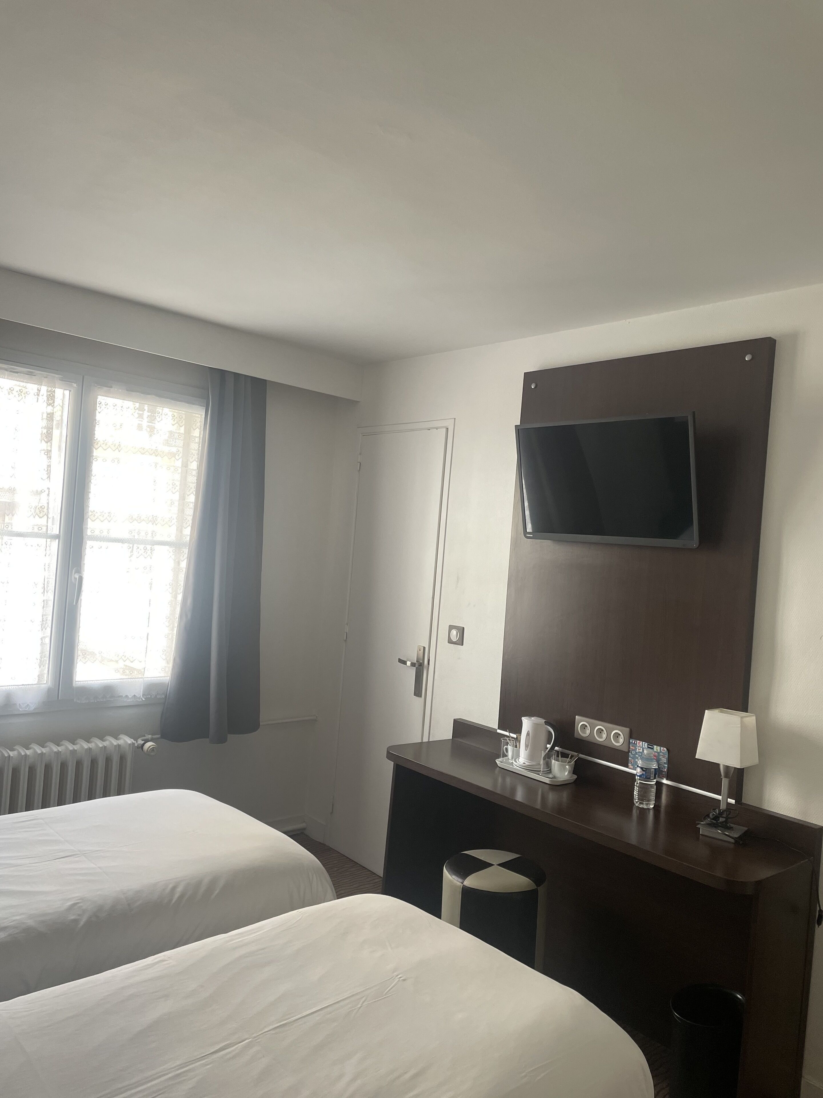 A hotel room with two beds, a desk with a television, a lamp, and a radiator.