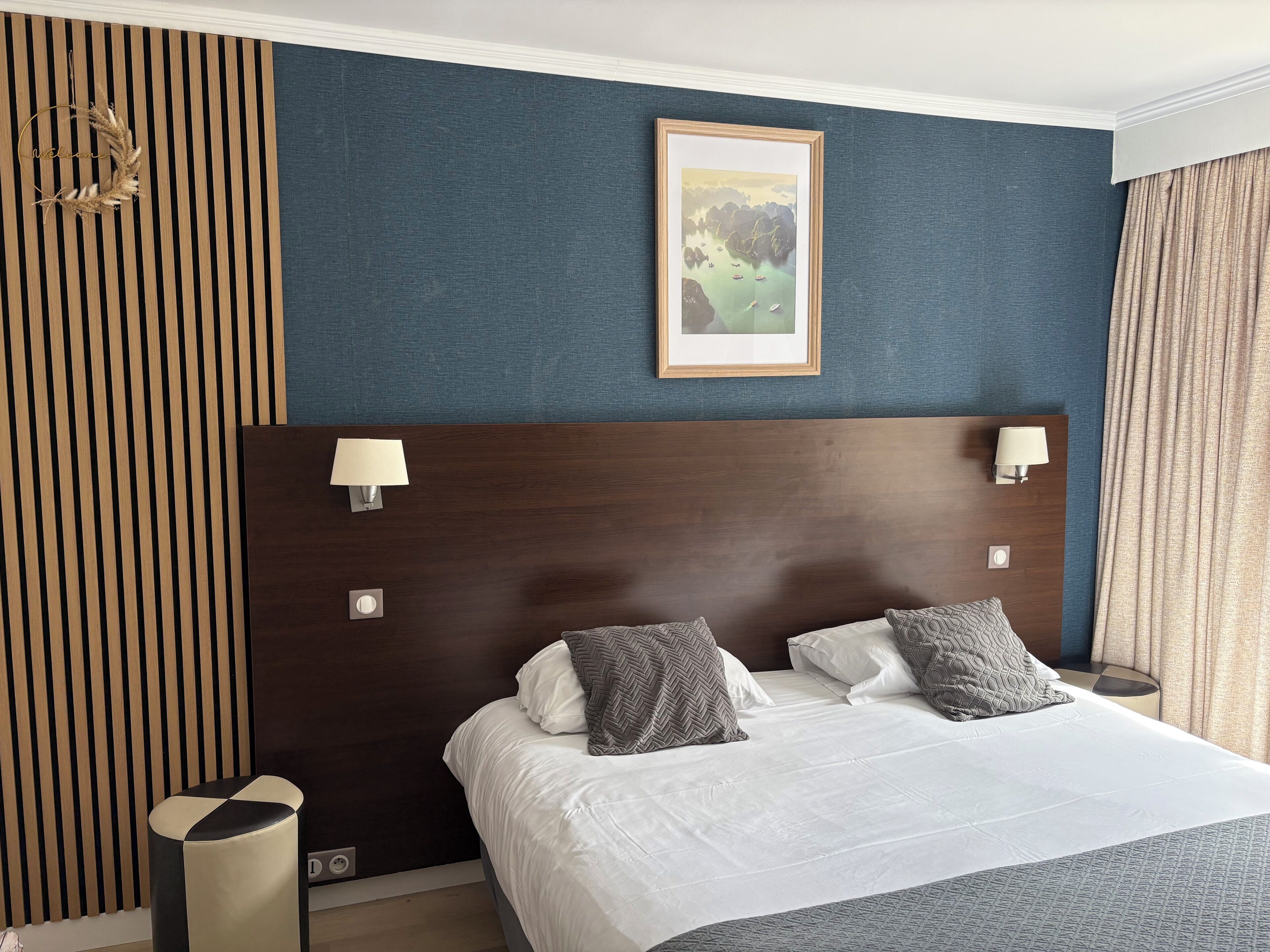 Habitación de hotel con cama, dos lámparas de noche, un cuadro en la pared y una ventana con cortinas.