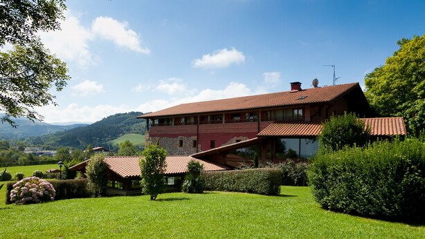 Une maison à deux étages, avec un toit rouge, entourée d’une pelouse et de haies bien entretenues, se dressant sur un fond de collines et sous un ciel dégagé.