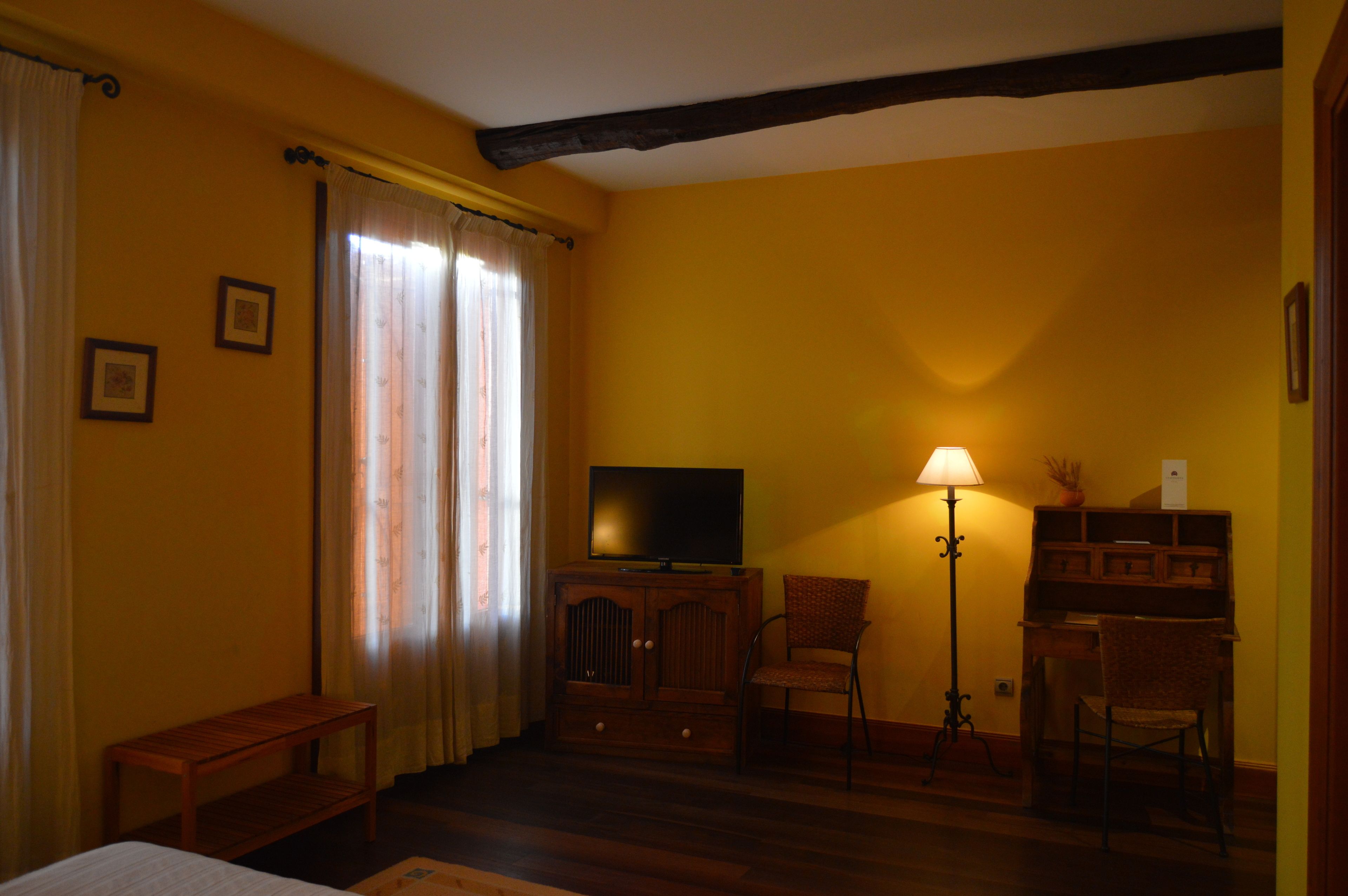Een kamer met gele wanden, een houten vloer, een televisie op een kast, een bureau met stoel, een lamp en een bankje.