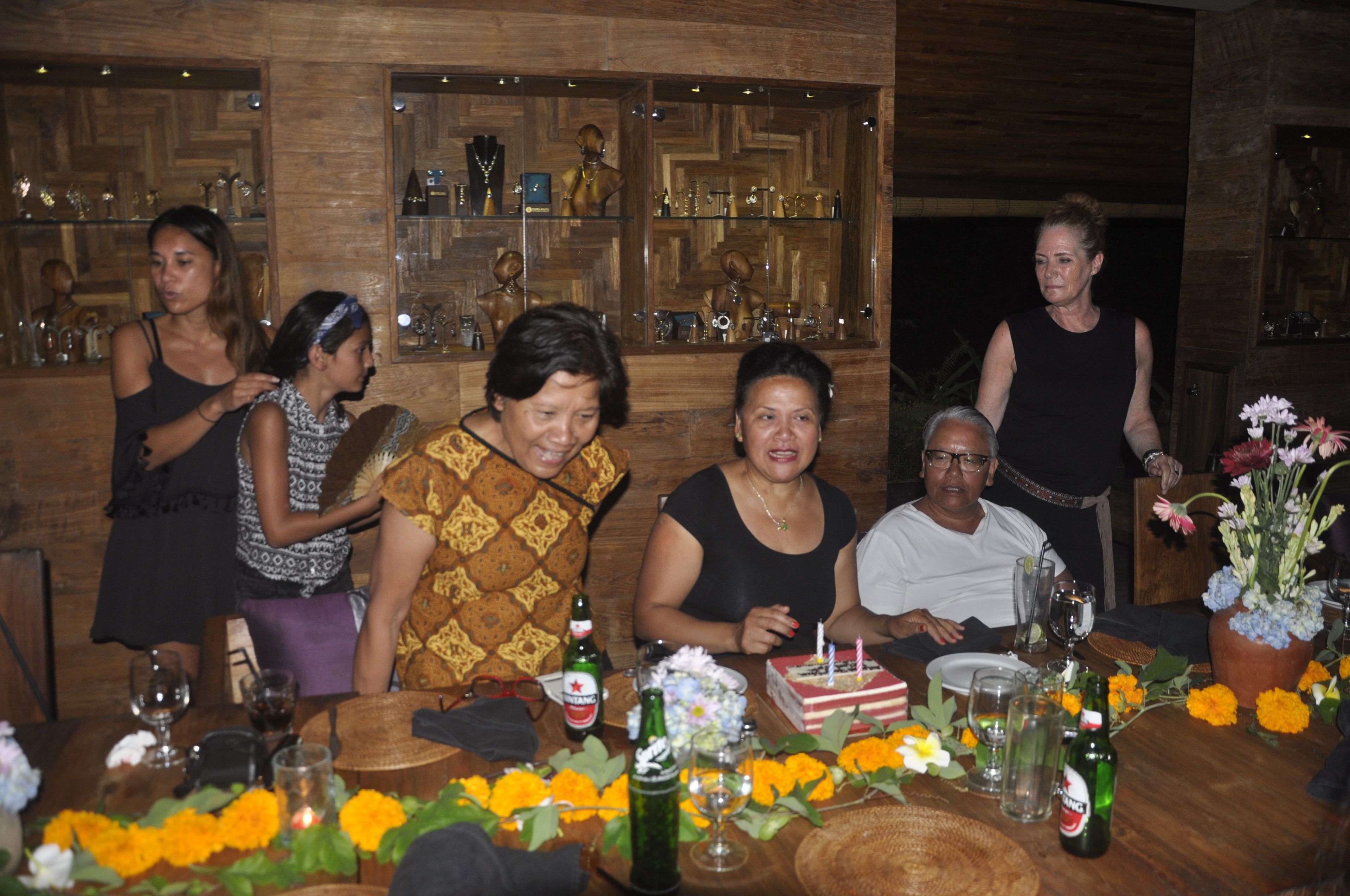 A group of people seated around a wooden table with a cake and drinks, in a room with wooden walls and shelves of glassware.