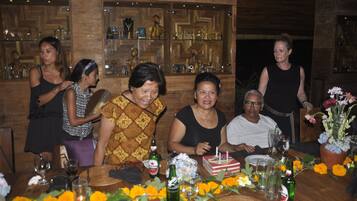 A group of people seated around a wooden table with a cake and drinks, in a room with wooden walls and shelves of glassware.