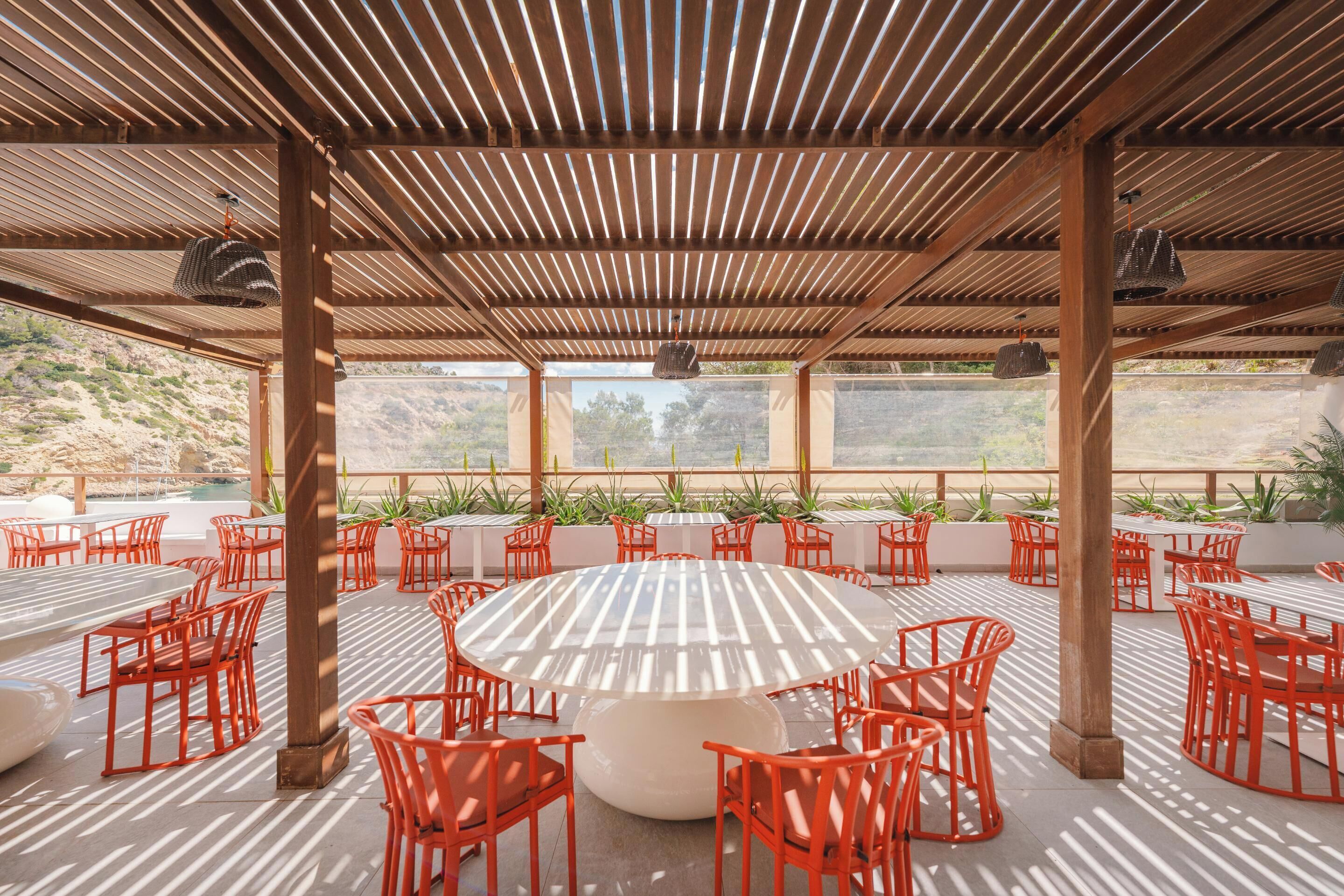 Une terrasse extérieure avec des tables rondes et des chaises orange, entourée de plantes en pot et offrant une vue sur un paysage pittoresque.