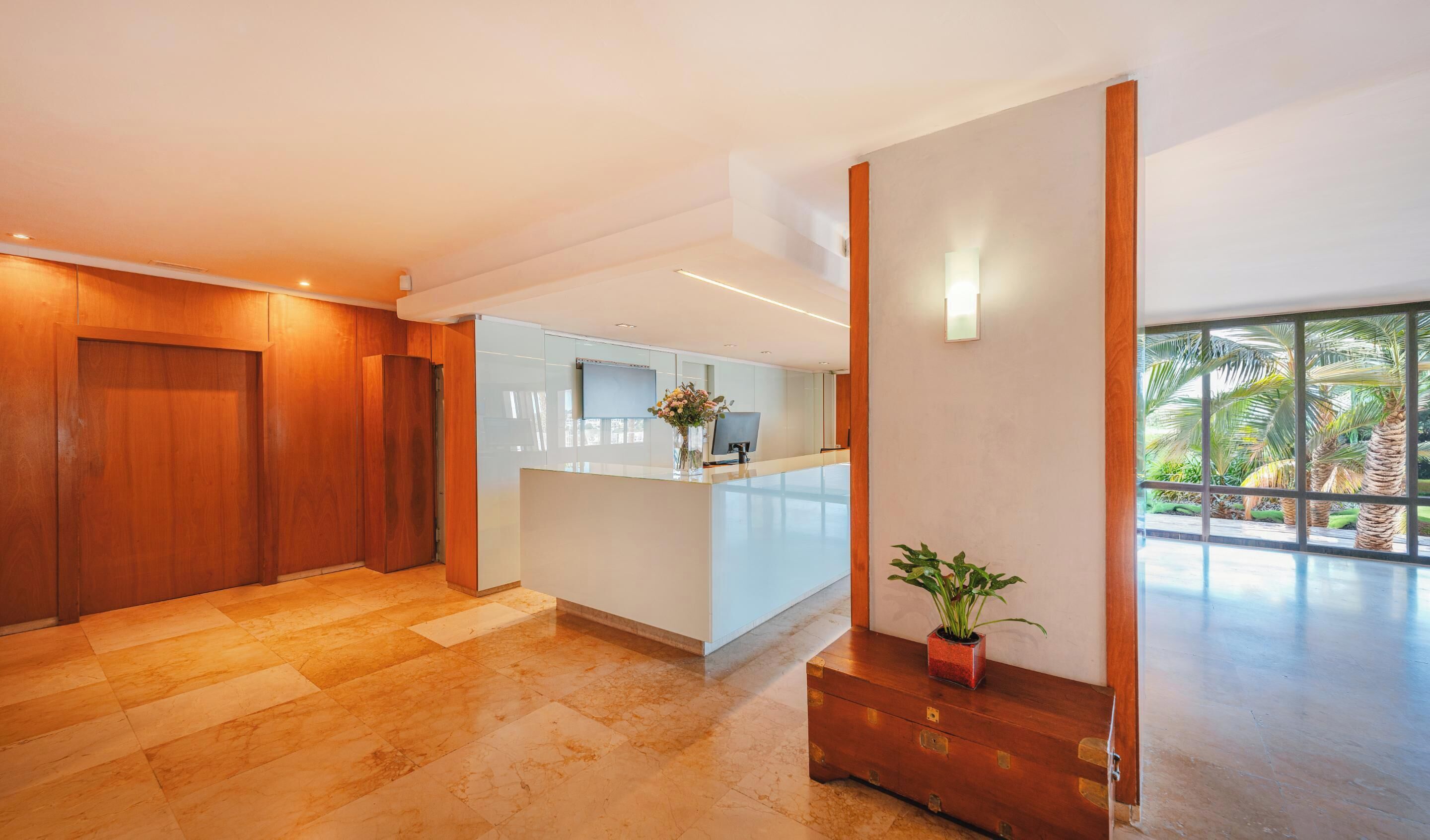 Eine moderne Hotelhalle mit einer Rezeption, einem Empfangsbereich aus Holz und einem großen Fenster mit Blick auf Palmen.