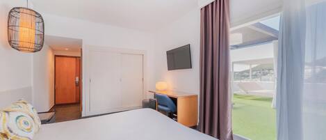 Habitación de hotel con una cama, un escritorio con lámpara, televisor y vistas a un paisaje verde.