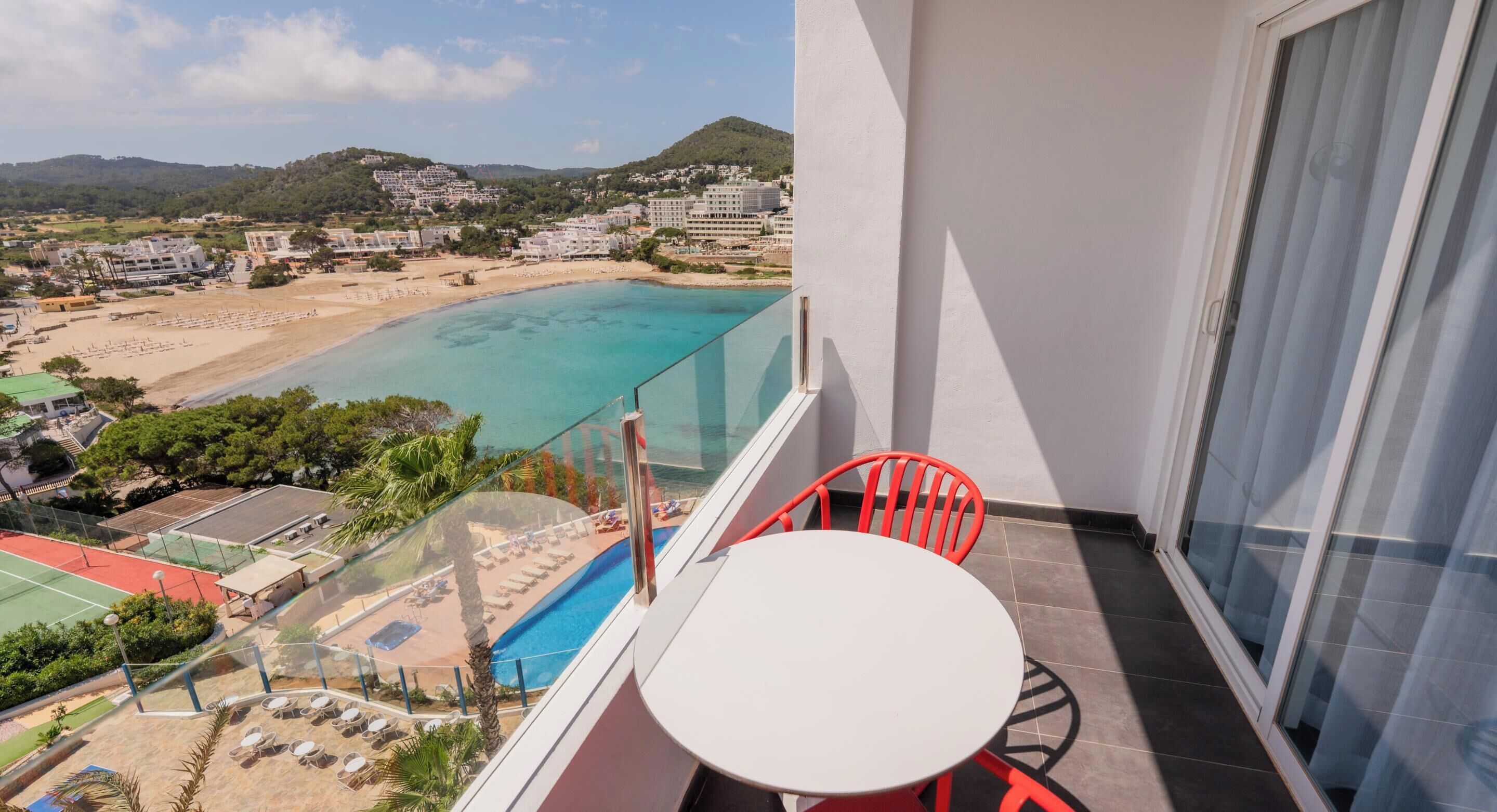 A balcony with a white table and red chairs, overlooking a beach and a pool.