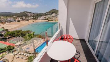 A balcony with a white table and red chairs, overlooking a beach and a pool.