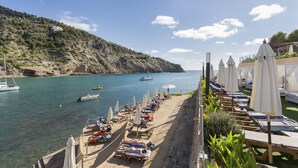 A beach with rows of sun loungers and umbrellas, a rocky hillside, and boats in the water. - Palladium Hotel Cala Llonga - Adults Only (Santa Eulalia del Rio)