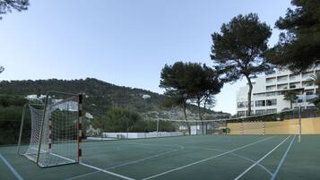 An outdoor tennis court with a goal post, surrounded by trees and a building in the background.