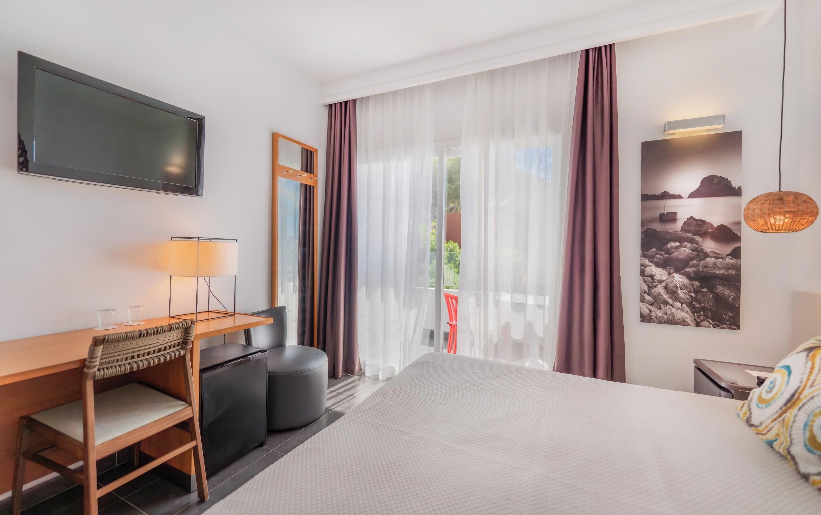 A hotel room with a bed, a desk with a chair, a television, a window with sheer curtains, and a wall-mounted picture.