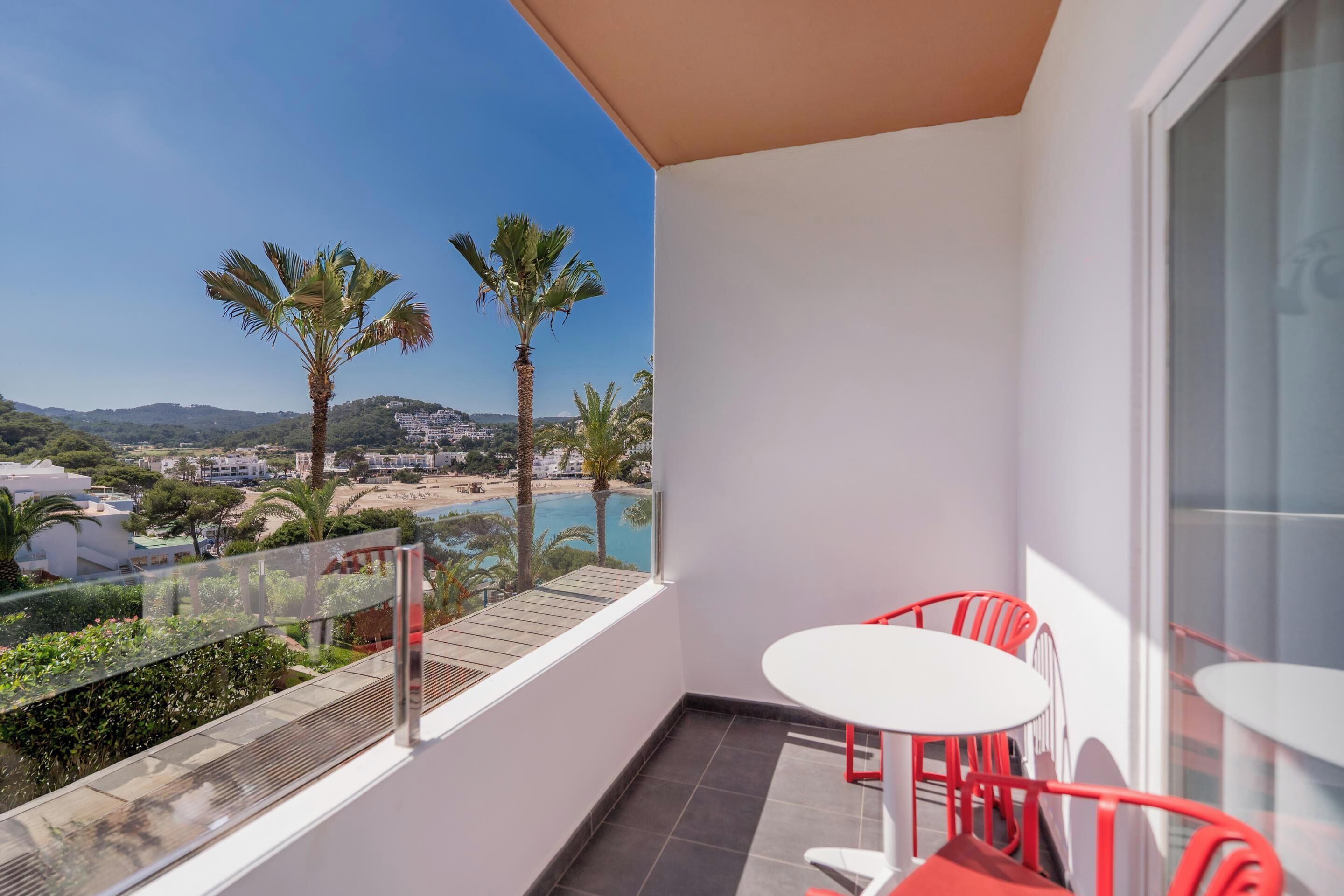 A balcony with a glass railing, a white table, and two red chairs.