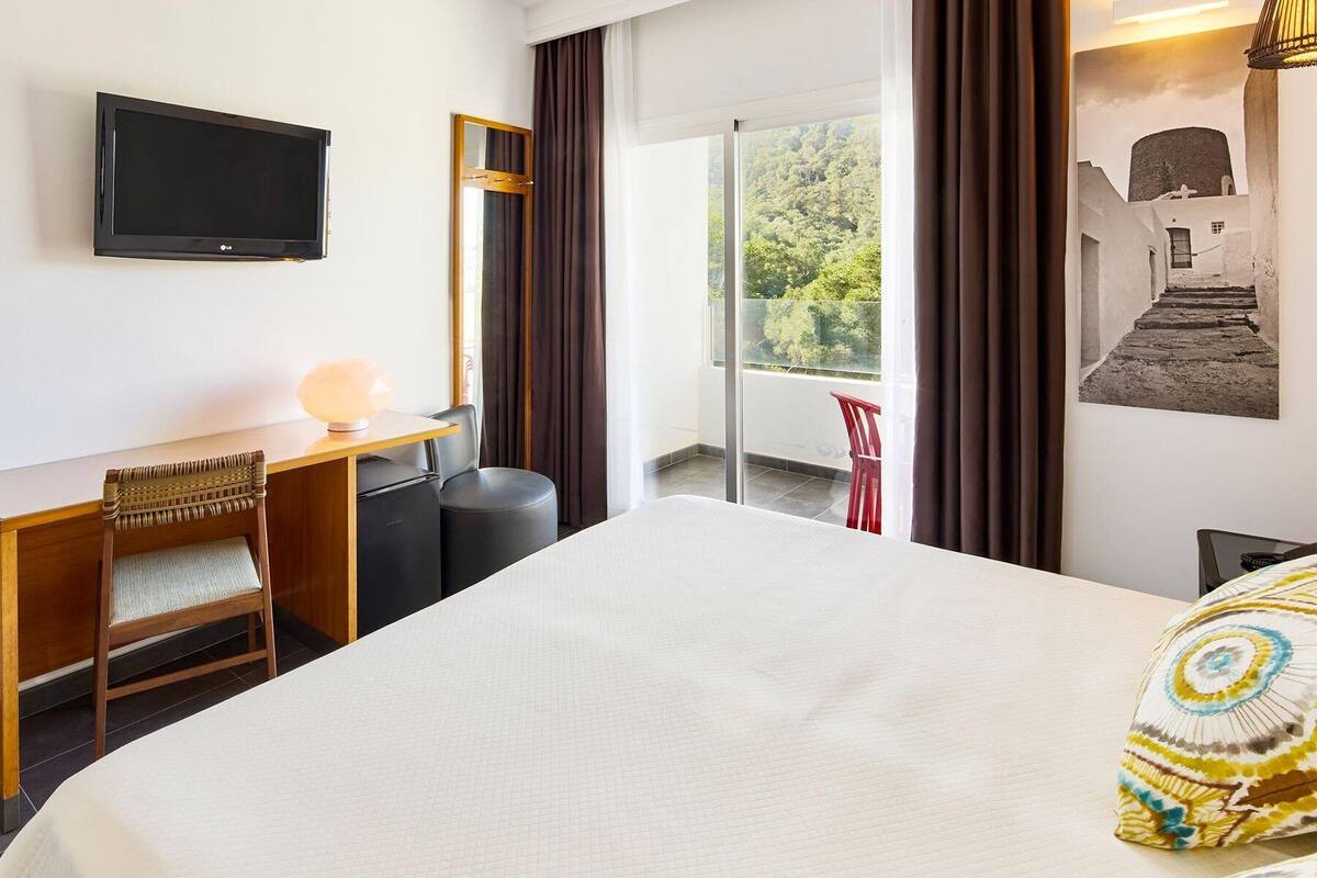 A hotel room with a bed, a desk with a chair, a television, a window with curtains, and a balcony with a chair.