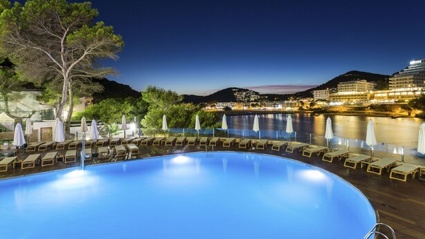 A swimming pool with lounge chairs and umbrellas, surrounded by a waterfront view and buildings in the background. - Palladium Hotel Cala Llonga - Adults Only (Santa Eulalia del Rio)