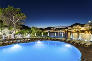 En swimmingpool med liggestole og parasoller, med udsigt til vandet og bygninger i baggrunden.