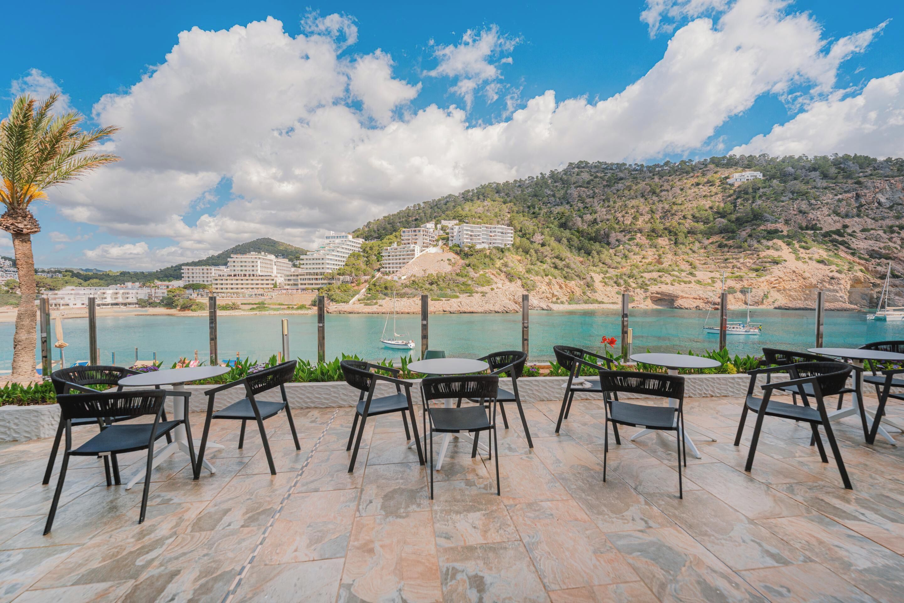 Un área de estar al aire libre con mesas y sillas, con vista a un paisaje costero con edificios y colinas.