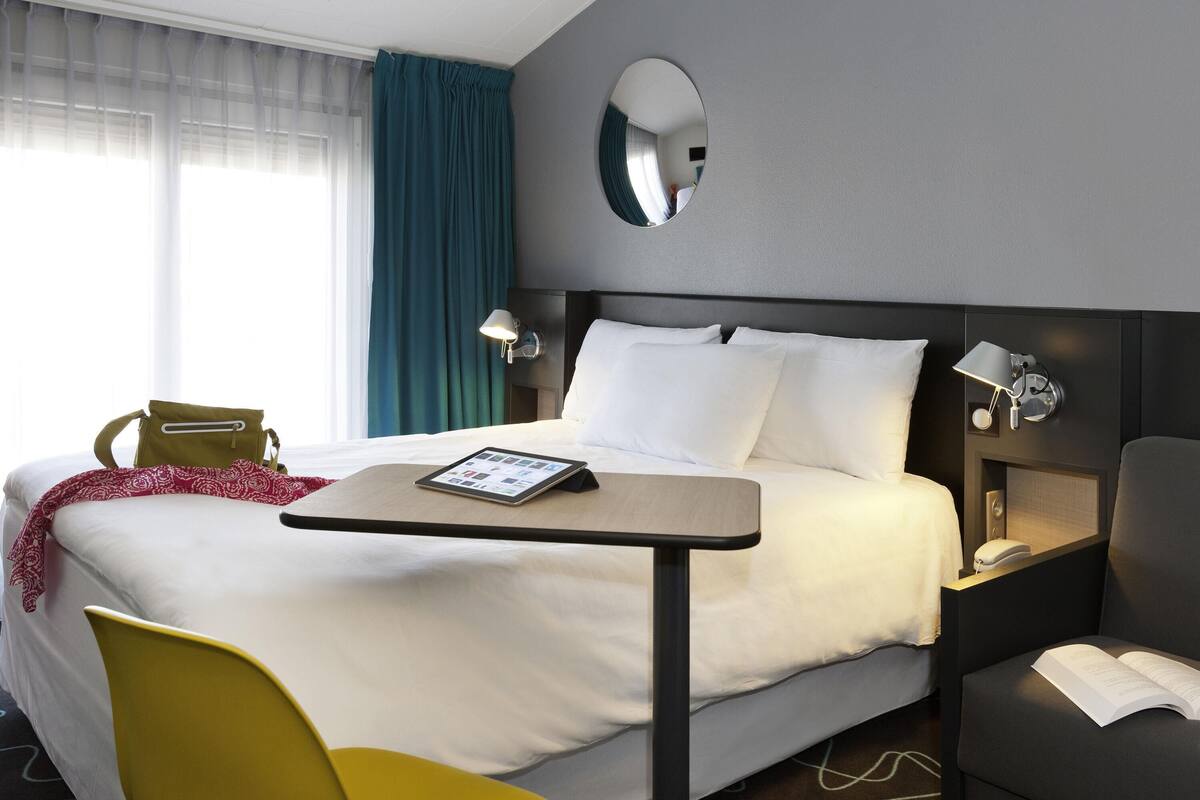 Habitación de hotel con cama, mesitas de noche, escritorio, silla, espejo y ventana con cortinas.