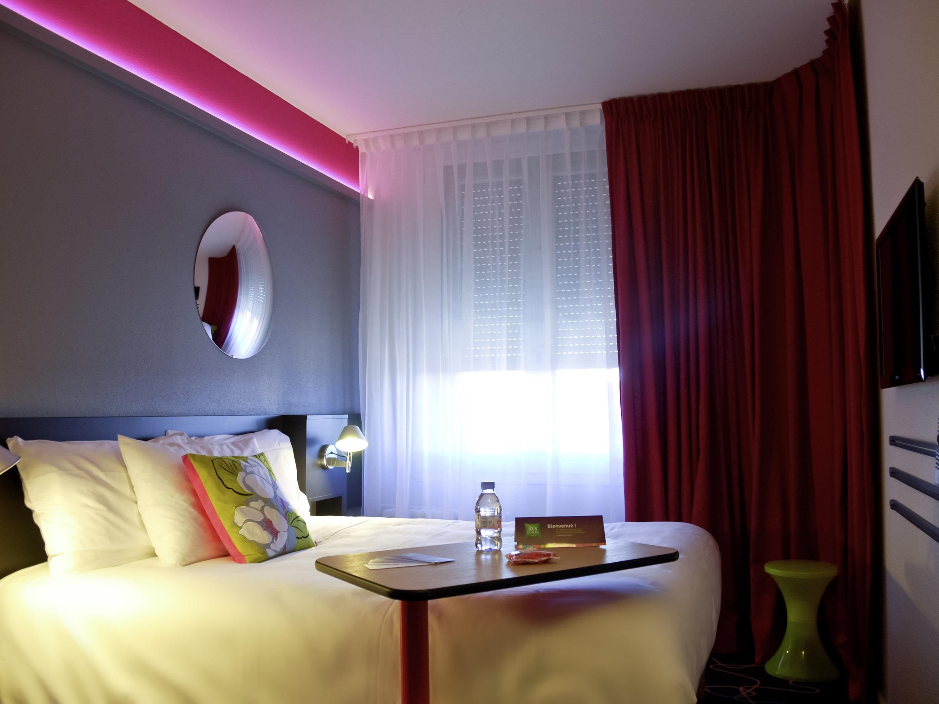 Une chambre d’hôtel avec un lit, une table de chevet sur laquelle se trouvent une bouteille et une carte, une lampe, un miroir et une fenêtre avec des rideaux.