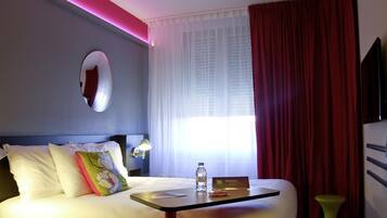 Une chambre d’hôtel avec un lit, une table de chevet sur laquelle se trouvent une bouteille et une carte, une lampe, un miroir et une fenêtre avec des rideaux.