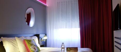 A hotel room with a bed, a bedside table with a bottle and a card, a lamp, a mirror, and a window with curtains.