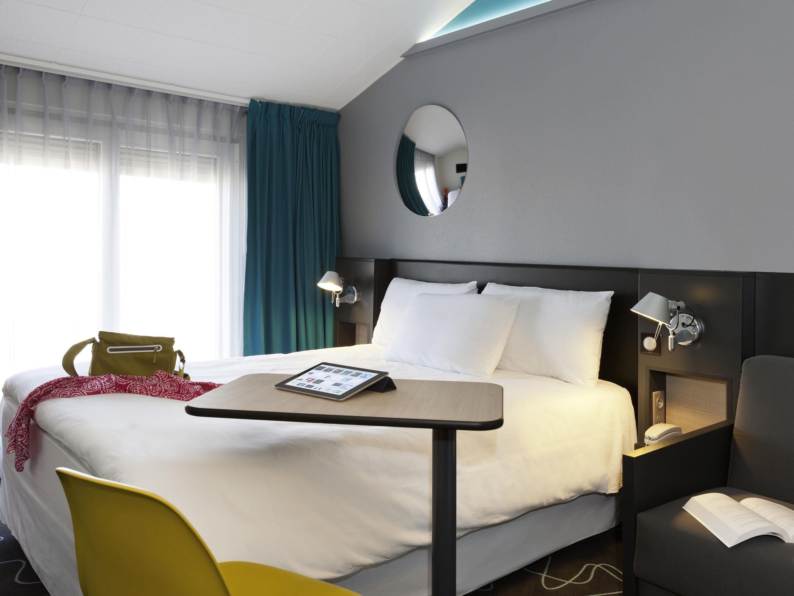 A hotel room with a bed, bedside tables, a desk, a chair, a mirror, and a window with curtains.